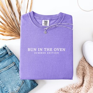 May include: A purple t-shirt with the text "Bun in the Oven Summer Edition" printed on the front.