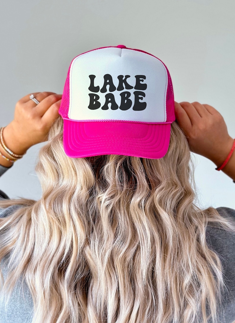 May include: Pink trucker hat with a white mesh back and black retro font that says "LAKE BABE".