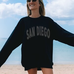 May include: A black sweatshirt with the text "SAN DIEGO" in a white outline on the front. The sweatshirt is being worn by a person standing on a beach.