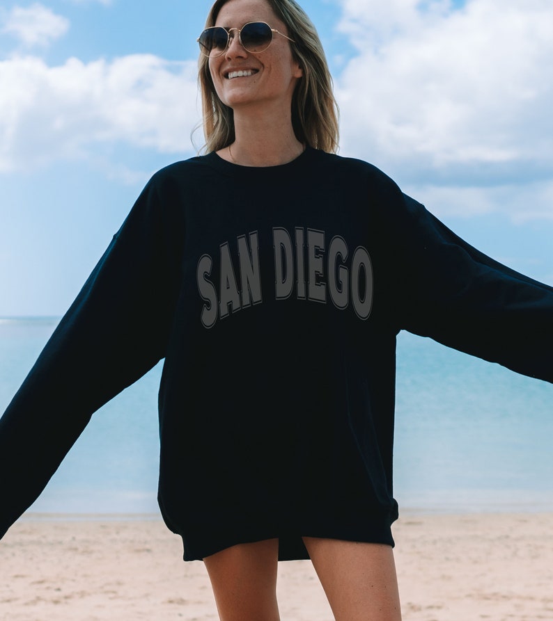 May include: A black sweatshirt with the text "SAN DIEGO" in a white outline on the front. The sweatshirt is being worn by a person standing on a beach.