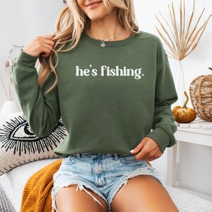 Puede incluir: Una sudadera verde con el texto "he's fishing." impreso en blanco. La sudadera la lleva puesta una persona que lleva pantalones cortos de mezclilla.