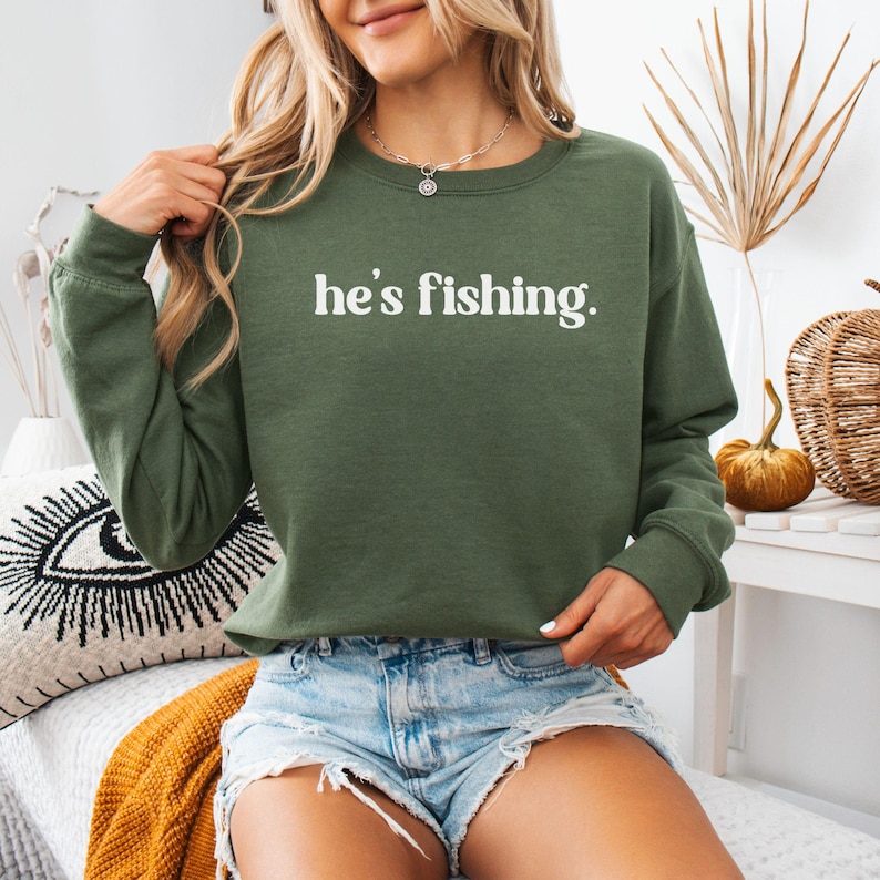 Puede incluir: Una sudadera verde con el texto "he's fishing." impreso en blanco. La sudadera la lleva puesta una persona que lleva pantalones cortos de mezclilla.