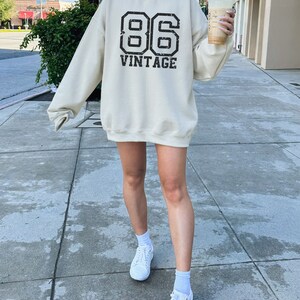 May include: A cream colored sweatshirt with a black vintage 86 graphic. The sweatshirt is oversized and has a crew neck.