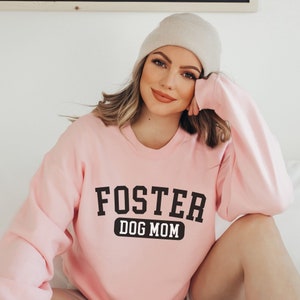 May include: A pink sweatshirt with the text "FOSTER DOG MOM" in black lettering. The sweatshirt has a crew neck and long sleeves.
