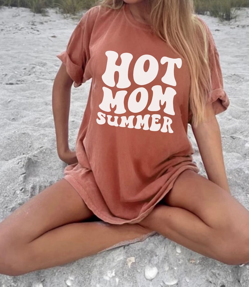 May include: A woman wearing a rust-colored t-shirt with the text "HOT MOM SUMMER" in white retro-style lettering. The shirt is oversized and has a relaxed fit.
