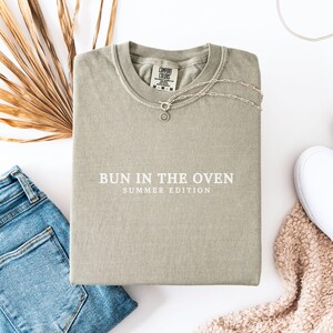 May include: A light green t-shirt with the text "Bun in the Oven Summer Edition" printed in white on the front.