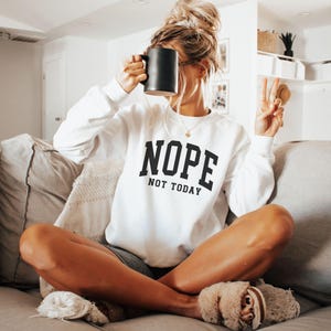 May include: White sweatshirt with black text that reads "NOPE NOT TODAY". A person is wearing the sweatshirt and holding a black mug.