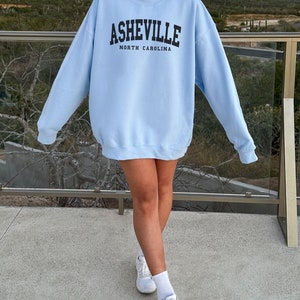 Pode incluir: Uma camisa de frio azul claro com o texto "Asheville, Carolina do Norte" estampado na frente.