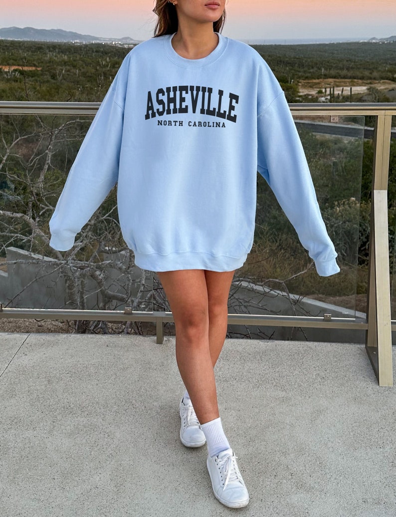 Pode incluir: Uma camisa de frio azul claro com o texto "Asheville, Carolina do Norte" estampado na frente.