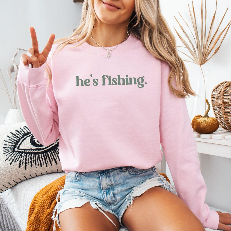 Puede incluir: Una sudadera rosa con el texto "he's fishing." en letras verdes.