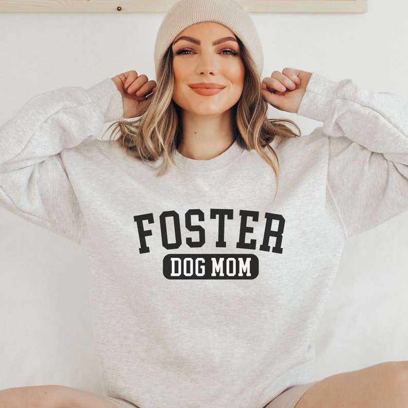 May include: A grey sweatshirt with the text "FOSTER DOG MOM" in black lettering. The sweatshirt is being worn by a person with blonde hair and a beige beanie.