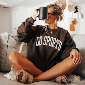 May include: A brown sweatshirt with the text "GO SPORTS" in white lettering. The sweatshirt is being worn by a person sitting on a sofa, holding a black mug.