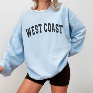 Puede incluir: Una sudadera de color azul claro con el texto "WEST COAST" impreso en negro en la parte delantera.