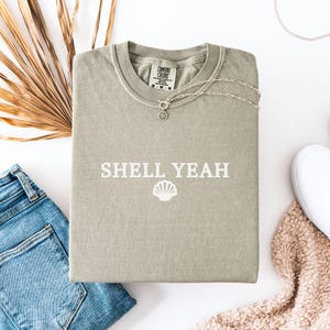 May include: A light green t-shirt with the text "SHELL YEAH" in white lettering and a white seashell graphic. The shirt is made of a soft, comfortable fabric.