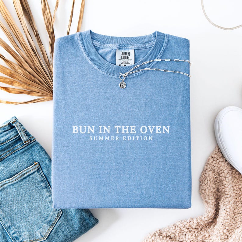 May include: A light blue t-shirt with the text "Bun in the Oven Summer Edition" printed in white.