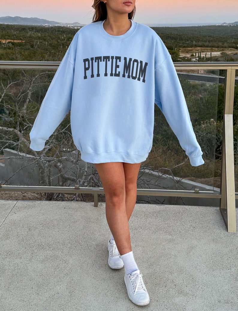 May include: A light blue sweatshirt with the text "PITTIE MOM" printed in black on the front. The sweatshirt is being worn by a person standing on a concrete surface.