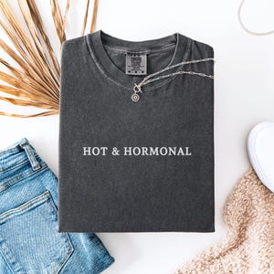 May include: A dark gray t-shirt with the text "HOT & HORMONAL" printed in white on the front. The shirt is folded and laid flat on a white surface.