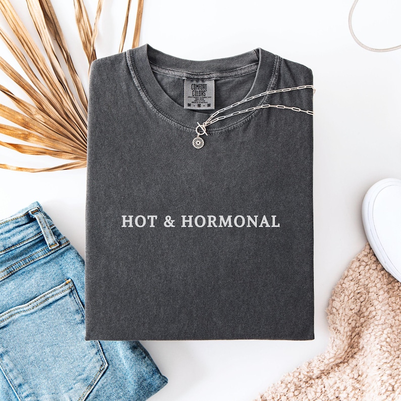 May include: A dark gray t-shirt with the text "HOT & HORMONAL" printed in white on the front. The shirt is folded and laid flat on a white surface.