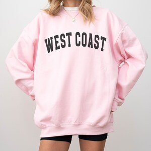 Puede incluir: Una sudadera rosa con el texto "WEST COAST" impreso en negro.