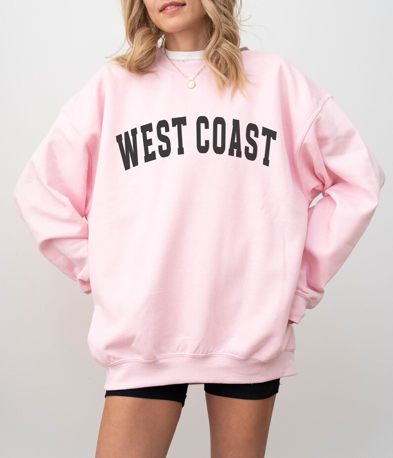 Puede incluir: Una sudadera rosa con el texto "WEST COAST" impreso en negro.