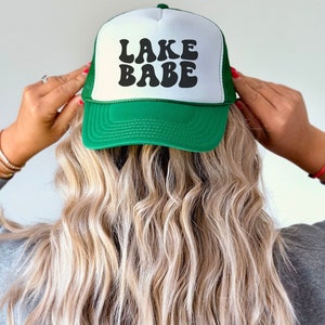 May include: A green and white trucker hat with the text "LAKE BABE" in black retro font.