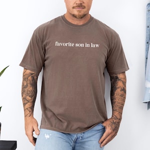 May include: A brown t-shirt with the text "favorite son in law" printed in white on the front.