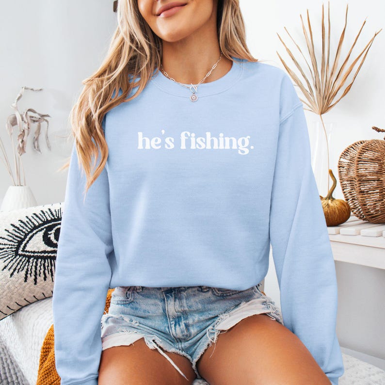 Puede incluir: Una sudadera de color azul claro con el texto "he's fishing." impreso en blanco.