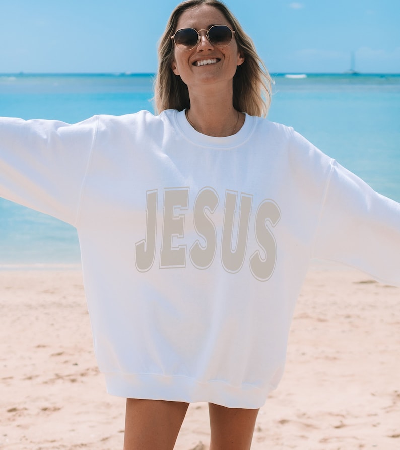 May include: White sweatshirt with the word "JESUS" printed in a beige color on the front. The sweatshirt is being worn by a person on a beach.