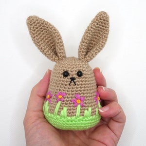 May include: A crocheted brown bunny with long ears, holding a green and pink flower pot with pink flowers.