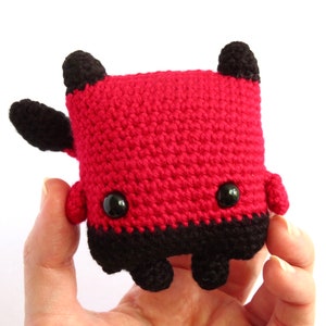 May include: A red and black crocheted plush toy with black eyes and horns. The toy is shaped like a square with rounded edges.