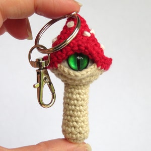 May include: A crocheted mushroom keychain with a red and white cap and a single green eye. The mushroom has a beige stem and is attached to a silver key ring.