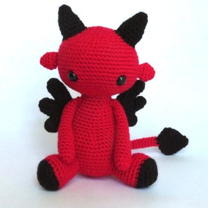 May include: A red crocheted devil plush toy with black horns, wings, and tail. The toy has black button eyes and is sitting on a white background.