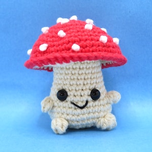 May include: A crocheted mushroom toy with a red cap and white spots. The mushroom has a white body with black eyes and a smiling mouth.