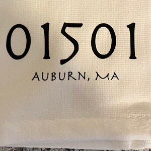 以下が含まれることがあります： 黒い文字で「01501 AUBURN, MA」と書かれた白いキッチンタオルです。