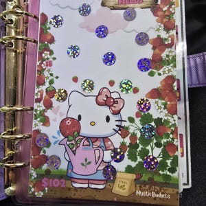 Może przedstawiać: Ilustracja różowej i białej Hello Kitty z konewką i rośliną truskawek. Ilustracja jest otoczona holograficznymi naklejkami z brokatem. Tekst "Scratch #s 3-7 STRAWBERRY FIELDS" znajduje się w górnej części obrazu. Tekst "6 €" i "102 €" znajduje się w dolnej części obrazu. Tekst "SEEDS" i "Mystic Budgets" znajduje się w prawym dolnym rogu obrazu. Tekst "Fits in your A6 Binder" znajduje się w dolnej części obrazu.