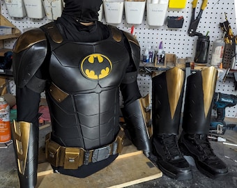 May include: A black and gold superhero costume, including a chest plate with a yellow bat symbol, matching arm and leg armor, and black boots. The costume is displayed on a mannequin, showcasing the details of the design.