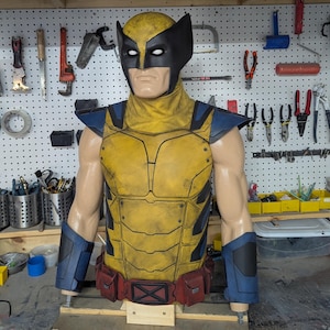 May include: A Wolverine costume torso piece featuring a yellow and black mask with white eye lenses, a yellow chest plate with black accents, blue shoulder and forearm guards, and a red utility belt with a black X-shaped buckle at the centre.