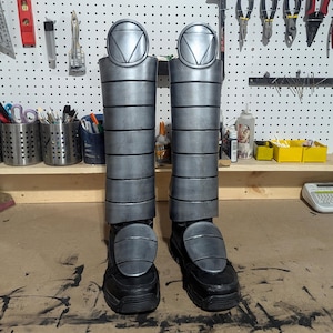 May include: A pair of silver and black boots with a geometric design on the front. The boots are made of a hard material and have a rounded toe.