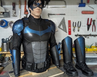 May include: A Nightwing costume displayed on a mannequin, featuring a black and blue chest piece, shoulder armour, forearm gauntlets, a utility belt, a blue domino mask, and a pair of black boots with matching blue shin guards for a complete set.