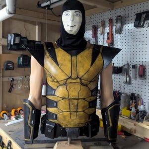 May include: A yellow and black costume with a black hood and a painted face. The costume includes a chest piece, shoulder pads, and arm guards. The chest piece has a detailed, textured design.