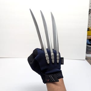 May include: A dark blue and black tactical glove featuring three long, silver-coloured, curved blades extending from the knuckles. The glove has a textured fabric construction with a small black tag on the side and a reinforced wrist strap closure.