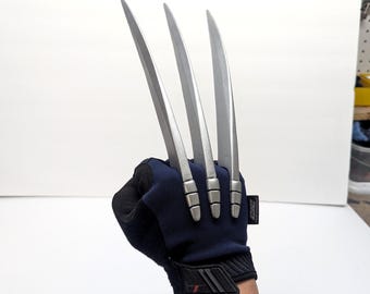 May include: A dark blue and black tactical glove featuring three long, silver-coloured, curved blades extending from the knuckles. The glove has a textured fabric construction with a small black tag on the side and a reinforced wrist strap closure.