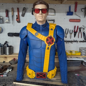May include: A blue and yellow costume, resembling a superhero uniform, is displayed on a mannequin. The costume features a yellow belt and shoulder straps, with a red "X" emblem on the chest. The mannequin wears red sunglasses.