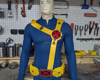 Può includere: Una maglia tattica blu a maniche lunghe con motivo testurizzato, dotata di tracolla e cintura portaoggetti gialle. Sia la tracolla che la cintura presentano un emblema a X rossa. Il busto è esposto su un manichino avvolto in nastro adesivo beige.