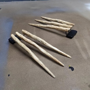 Puede incluir: Conjunto de seis garras de madera, pintadas en un color marrón claro, con una superficie rugosa y texturizada. Las garras están dispuestas en dos grupos, con tres garras en cada grupo. Las garras están montadas sobre una base negra.