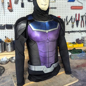May include: A black and purple superhero costume with a silver belt. The chest piece is purple with black accents and a blue upper section. The costume includes shoulder armor and a black hood.