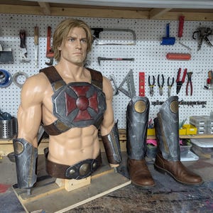 May include: A male mannequin torso wearing a brown leather chest harness with a red cross emblem, brown leather belt, and metallic arm guards, displayed alongside a pair of brown leather boots with metallic shin guards attached to the front.