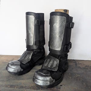 May include: Pair of black boots with silver-colored armor plating on the shins, ankles, and toes. The boots have adjustable straps and buckles. The boots are designed for protection and have a rugged, industrial aesthetic.