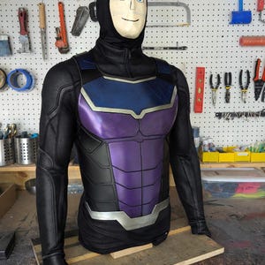 May include: A black and purple superhero costume with a blue chest plate. The costume has a black hood and long sleeves. The chest plate is purple with silver trim. The costume is displayed on a mannequin.
