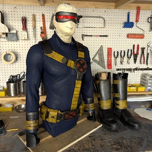 Può includere: Un costume da supereroe blu e giallo, tra cui tuta, cintura e stivali, esposto su un manichino. Il costume presenta una visiera rossa e un'emblema "X". Lo sfondo mostra un'officina con attrezzi.